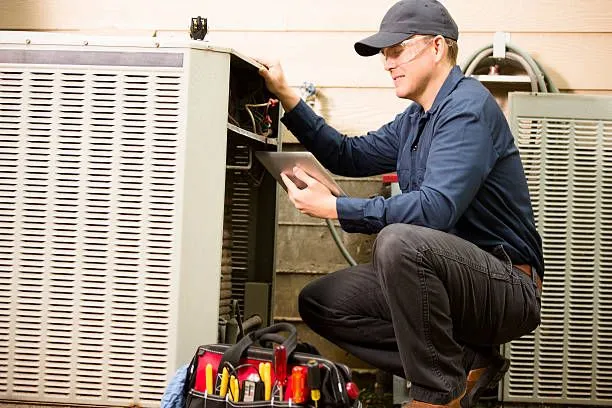 An AC technician is squatting next to an outdoor AC unit, performing repairs.