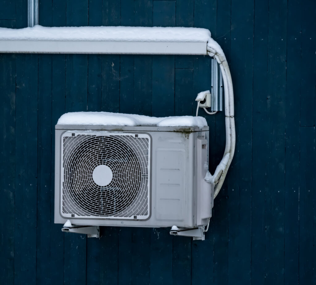 Heat pump mounted on the wall of a house and covered in snow on a cold winter day.